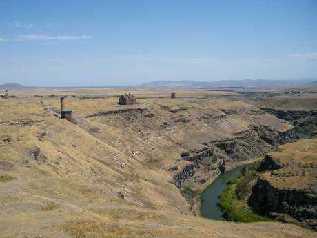 A wide view of the Ani Ruins, with multiple ancient structures spread across a vast, arid landscape overlooking the river that separates Turkey and Armenia.