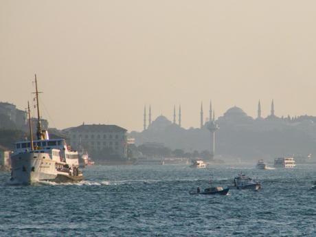 Boats sailing in the Bosphorus with a hazy view of Istanbul in the background, featuring the silhouette of famous mosques with their minarets piercing the sky.