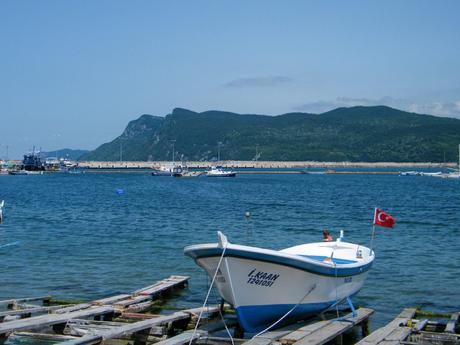 A small fishing boat with a Turkish flag docked near a wooden pier, with a backdrop of lush green hills and a clear blue sky.