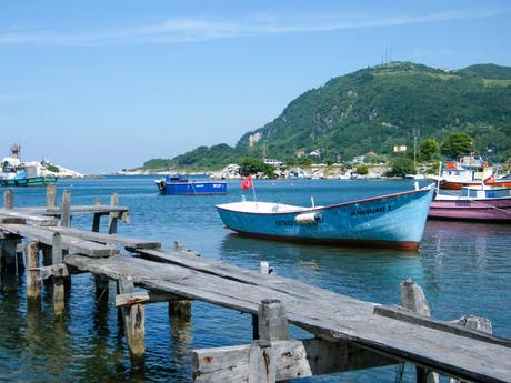 A turquoise fishing boat moored beside a wooden pier in a calm bay, with other colorful boats in the background and green hills in the distance.