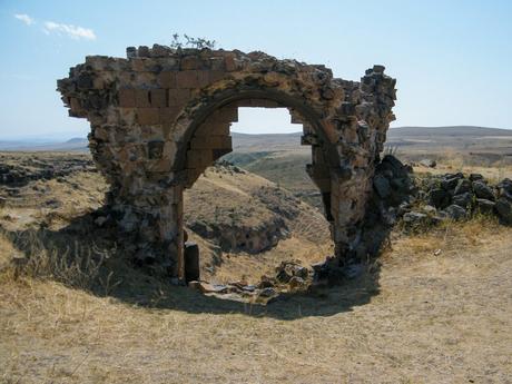 A crumbling stone archway standing alone in a dry, grassy landscape, with distant hills visible in the background.