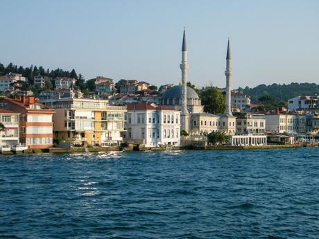 A waterfront view in Istanbul, Turkey, showing a mix of modern and traditional buildings, including a mosque with twin minarets, against a backdrop of a wooded hill.