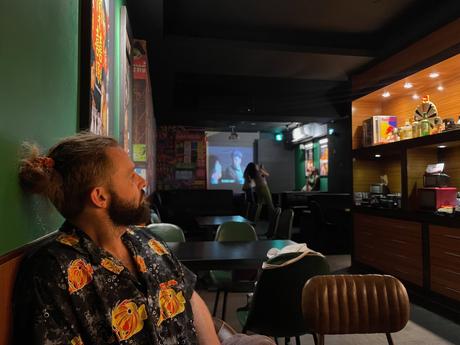 A cozy bar interior with dim lighting, Alex Tiffany wearing a floral shirt seated, and a film being projected on a screen in the background. Shelves display bottles and memorabilia.