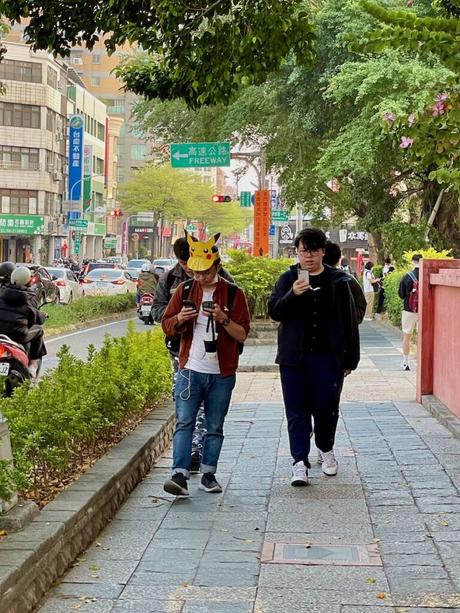 Two pedestrians walking along a tree-lined urban street, one wearing a bright yellow Pikachu hat, with shops, traffic signs, and vehicles in the background.