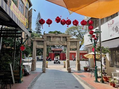 A stone archway at the end of Fuzhong Street, Tainan, decorated with red lanterns, opening onto a street with a temple in the background, under a bright sunny sky.