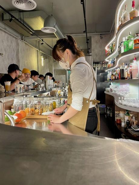 A bartender in a modern cocktail bar in Tainan preparing a drink at a polished counter surrounded by jars of ingredients, with shelves of liquor bottles lit in soft lighting behind her.