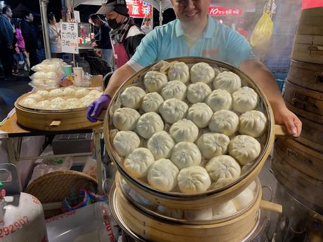 A vendor at a night market holding a steaming tray of freshly made buns, with additional stacks of bamboo steamers and a busy background of market stalls.