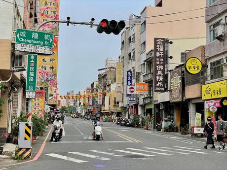 A buzzing Taiwanese street lined with shops and colorful signs in both Chinese and English, with scooters and pedestrians populating the road.