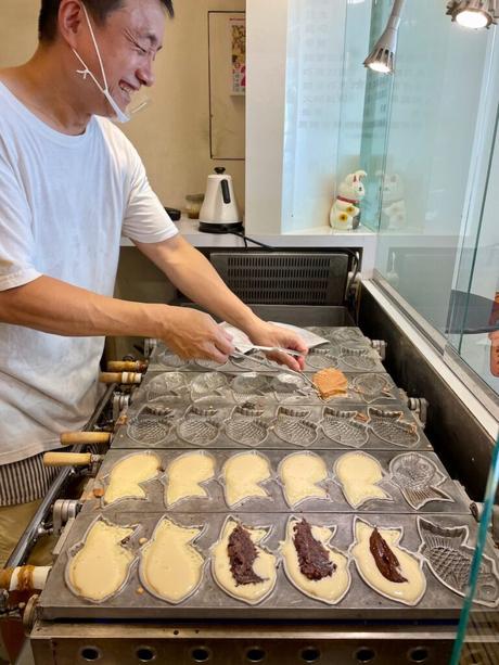 A smiling man wearing a white shirt as he cooks taiyaki-shaped pastries on a hot mold, with fillings like red bean paste and chocolate visible in the batter.