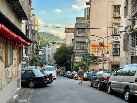 A quiet residential street in Taipei lined with cars, scooters, and buildings, with red lanterns hanging along a wall and mountains visible in the background.