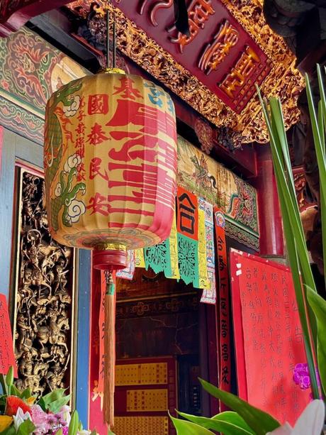 A red and gold lantern with intricate Chinese characters hanging inside a temple, surrounded by vibrant red pillars and detailed carvings.