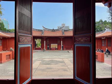 A view through an open red doorway into the main courtyard of the traditional Chinese Confucian Temple in Tainan with red walls, sloping roofs, and a few people walking or sitting under the bright sky.