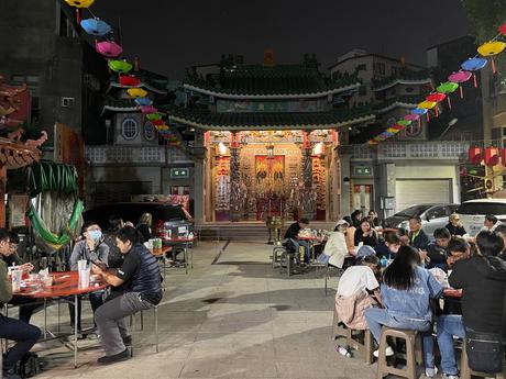 A vibrant temple scene at night, with colorful lanterns hanging overhead and diners enjoying meals at outdoor tables near a richly decorated temple facade.