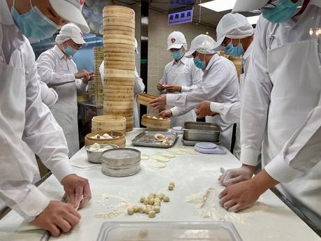 Chefs in white uniforms and masks at Din Tai Fung working together to prepare dumplings in a kitchen, with bamboo steamers and flour-covered counters.