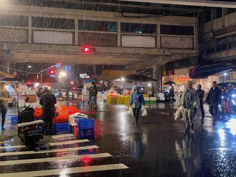 A busy fish market under an overpass, with people walking through the rain, carrying bags, and vendors operating stalls surrounded by crates and containers.