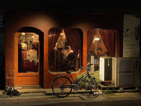 A cozy, warmly-lit cafe with wooden decor, red curtains, and a bicycle parked outside, offering a glimpse of people dining inside through large glass windows.