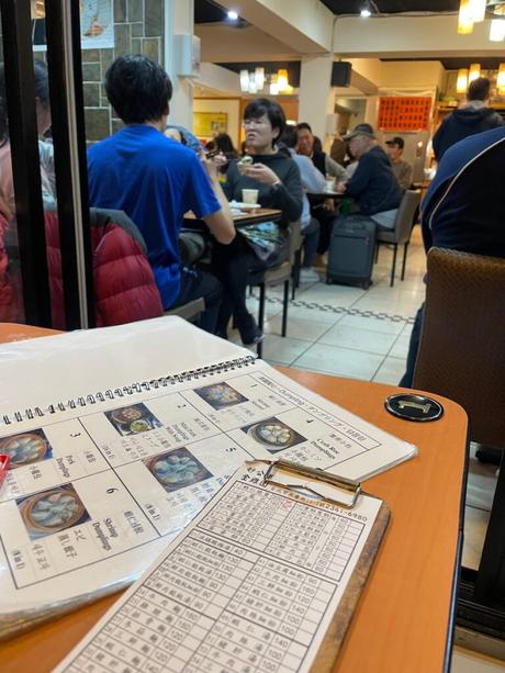 An indoor restaurant scene with tables filled with diners, a laminated menu in the foreground showing a selection of dumplings and dishes.