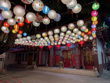 A vibrant display of colorful lanterns hanging from strings above the entrance of a traditional temple at night, with intricate red and gold door carvings in the background.