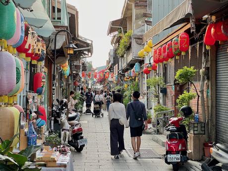 A lively alleyway in Taiwan with colorful paper lanterns hanging above, boutique shops, and visitors strolling. Scooters and decorative displays line the path.