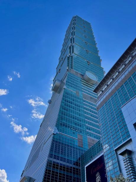 A dramatic upward view of Taipei 101, a tall, iconic green glass skyscraper against a bright blue sky with scattered clouds.