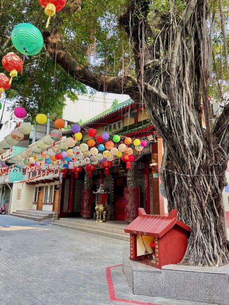 A temple courtyard during the daytime, adorned with bright multicolored lanterns hanging from the branches of a large tree, with a red altar and golden incense burner at the entrance.