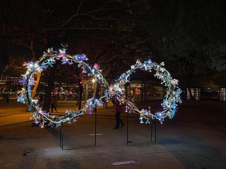 A decorative infinity loop sculpture illuminated with colorful lights at night, surrounded by a darkened park and faintly visible visitors.