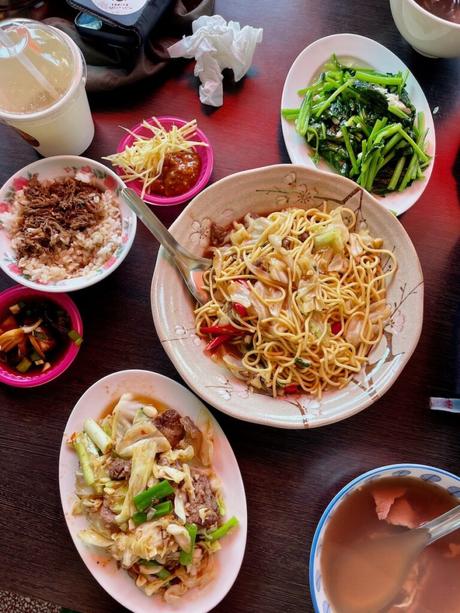 A close-up of a colourful Taiwanese meal featuring stir-fried noodles, rice with shredded meat, sautéed greens, and a selection of side dishes on a wooden table.