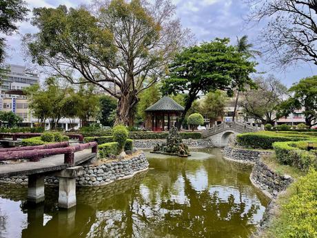 A serene garden with a pond, stone bridges, lush greenery, and a small pavilion surrounded by manicured bushes and trees.