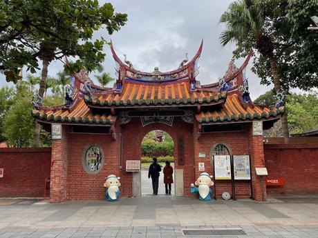 The arched entrance of a temple with a traditional tiled roof, flanked by statues and decorated with colorful carvings, leading into a courtyard.