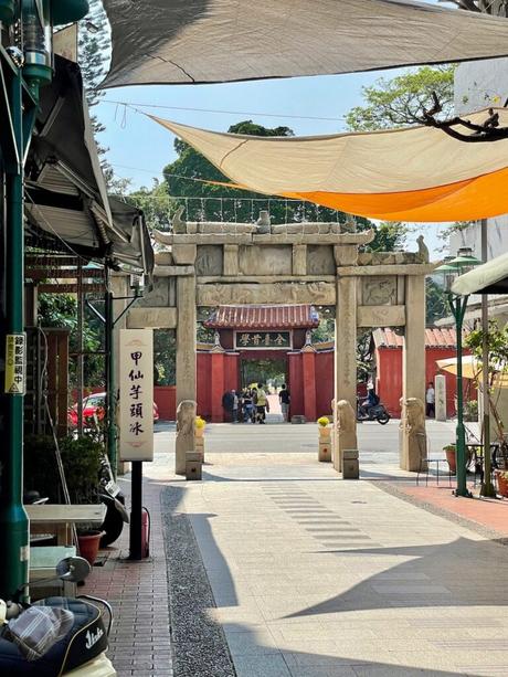 A scenic gateway in a traditional Taiwanese style, framed by a stone arch and flanked by small lion statues, leading to a red temple in the distance.