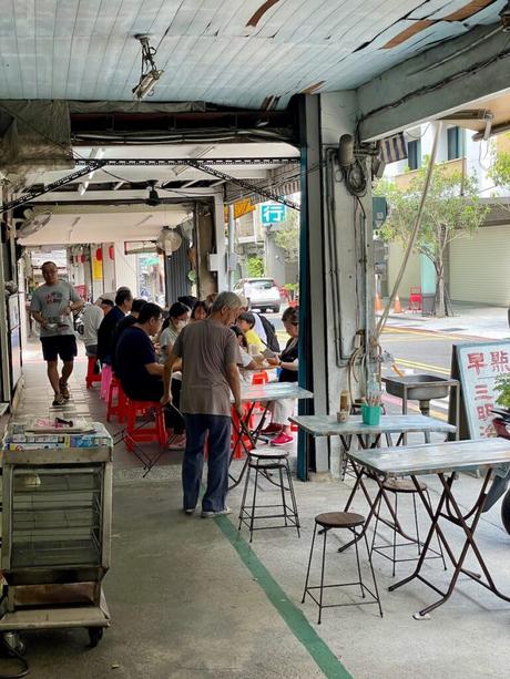 A casual outdoor dining area under a rustic canopy, with patrons enjoying meals at simple tables and a man walking past in the foreground.