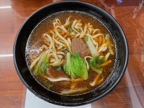 A steaming bowl of Taiwanese beef noodle soup with tender chunks of beef, bok choy, and thick noodles, served in a black bowl on a wooden table.