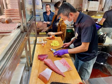 A fishmonger wearing gloves and a mask cuts fresh fish on a wooden counter, with various cuts of fish and seafood displayed in a busy market setting.