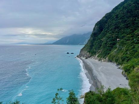 A stunning view of a turquoise ocean meeting a sandy beach, bordered by towering green cliffs under an overcast sky.