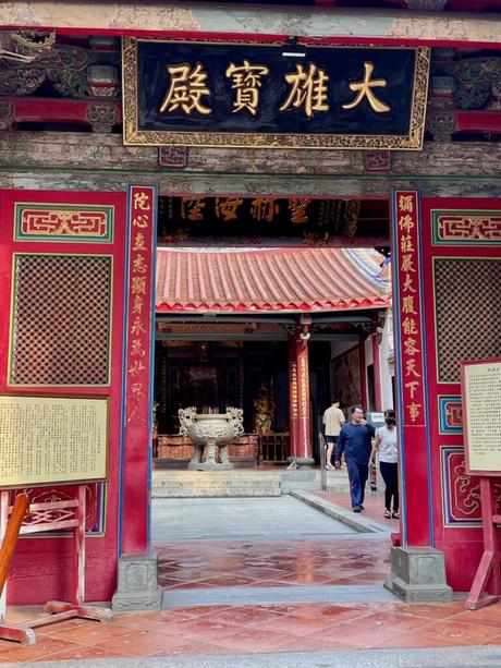 The entrance to a historic Taiwanese temple with bold red pillars, golden Chinese inscriptions, and intricate carvings. Visitors are seen exploring the interior courtyard.