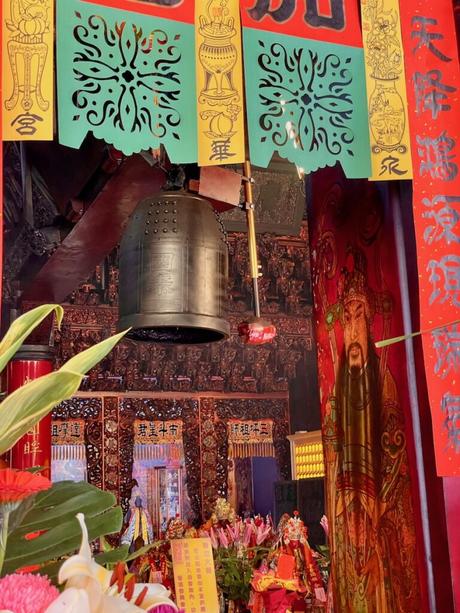 A detailed close-up of a traditional temple altar, featuring intricate wooden carvings, a large hanging bell, and colorful banners with Chinese characters.