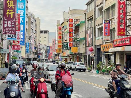 A busy intersection with people riding scooters and motorbikes, flanked by buildings with vibrant store signs and advertisements in Chinese characters.