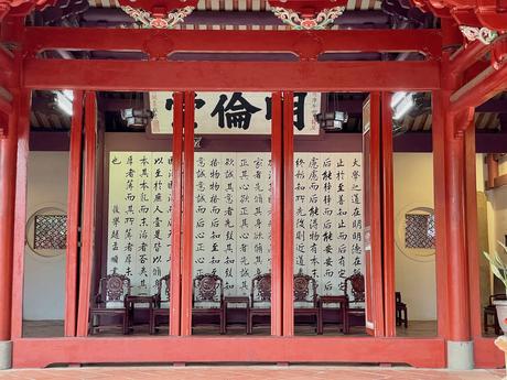 A traditional Chinese temple interior with vibrant red pillars, decorative carvings, and walls featuring large calligraphy panels, along with a row of antique wooden chairs.