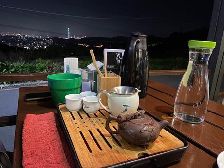 A wooden table with a traditional Chinese tea set, including teapots and cups, overlooking a cityscape lit up at night in the background.