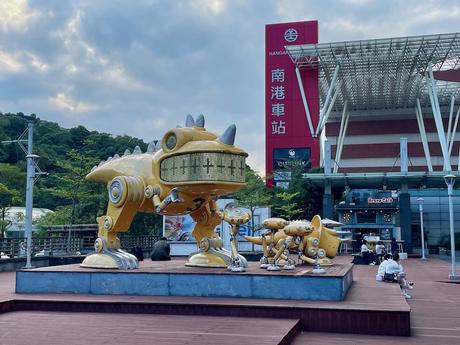 A set of large, metallic robotic creatures resembling dinosaurs, displayed outdoors near Nangang train station, with modern architecture in the background.