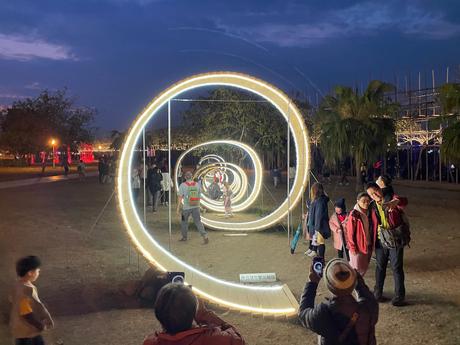 A series of glowing light rings forming a tunnel-like installation at night, with visitors walking through and taking photos under the dark blue sky.