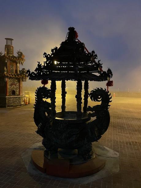 A decorative incense burner with intricate dragon motifs silhouetted against a foggy evening, illuminated by a soft yellow light in the background.