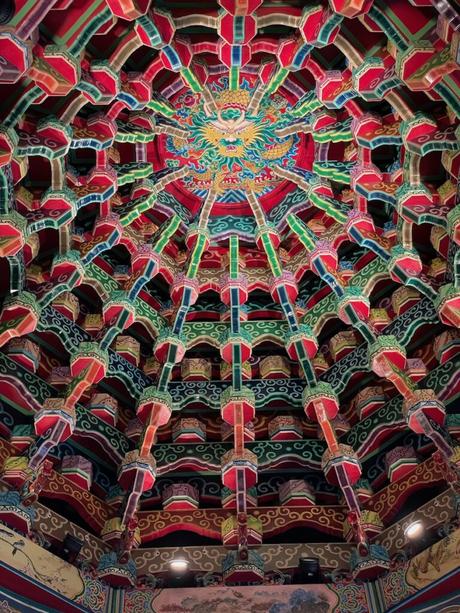 An elaborate and vividly painted temple ceiling featuring a detailed dragon motif surrounded by geometric designs in vibrant reds, greens, and golds.