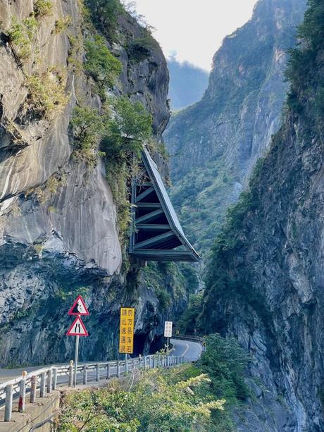 A winding mountain road with a protective steel overhang attached to the steep cliff face, surrounded by rugged rocky walls and dense greenery.