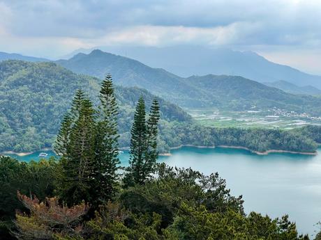 A stunning landscape with pine trees framing a vibrant turquoise lake, rolling hills, and distant villages, under a dramatic cloudy sky.