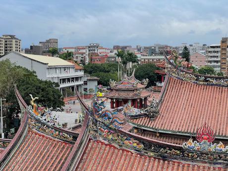 A close-up of intricately decorated temple rooftops featuring dragon and phoenix sculptures in vibrant colors, against a backdrop of trees and urban buildings.