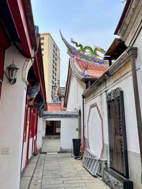 A narrow alleyway leading to a temple, with ornate roof designs featuring a colorful dragon sculpture, surrounded by white walls and red door frames.