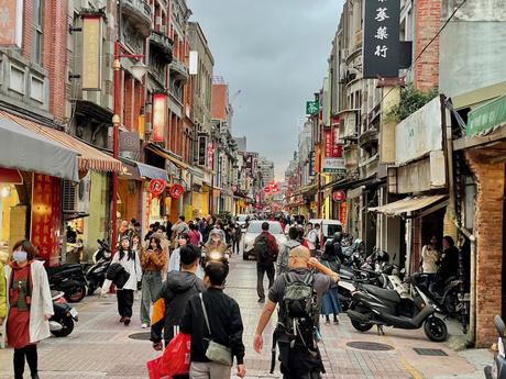 A bustling street market filled with people walking and shopping, surrounded by red-brick buildings with traditional Chinese signage, scooters parked along the sides.