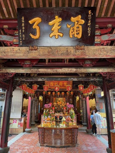 An ornately decorated altar in a Taiwanese temple, featuring vibrant offerings, intricate carvings, hanging red lanterns, and a sign with golden Chinese characters.