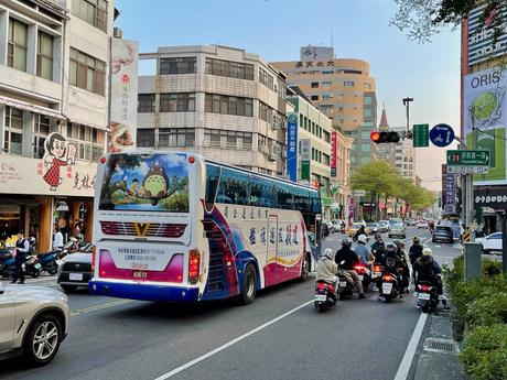 A colorful Taiwanese city street with a bus featuring Totoro-themed artwork on its back, surrounded by scooters and urban buildings under a clear sky.